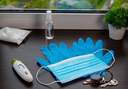 Medical mask, blue disposable gloves, thermometer, antiseptic gel, antibacterial wipes and house keys on the windowsillの写真素材