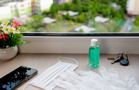 Disposable white medical gloves, mask, antiseptic gel, smartphone and house keys on the windowsillの写真素材