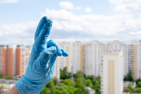Male hand in medical glove shows fingers crossed, superstition and lucky gesture against a cityscapeの写真素材