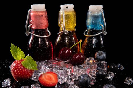 Strawberries, blueberries, cherries, mint leaves, ice cubes and bottles with strawberry, blueberry and cherry juice with drops of water on a black background with reflectionの写真素材