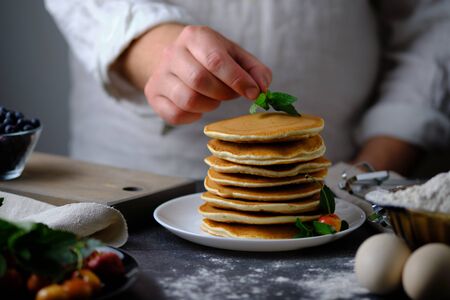 Man decorates pancakes with mint. On the table are the products from which they prepared the dish and berries. Dad prepares a delicious breakfast. The hands of the cook in the frame.の写真素材