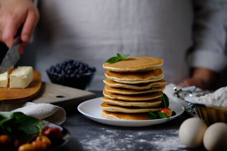 A man cuts off a piece of butter to decorate delicious pancakes. On the table are the products from which they prepared the dish and berries. Dad prepares a delicious breakfast. The hands of the cook in the frame.の写真素材