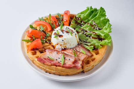 Hearty Belgian waffles with poached egg, bacon, lettuce, tomato and arugula, garnished with sesame seeds and green onions. Dish on a white plate.の写真素材