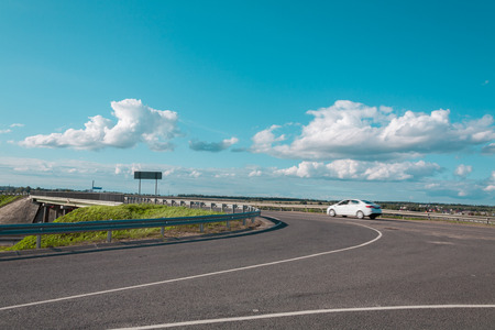 the white car at a speed passes a road fork against the blue sky and fluffy cloudsの写真素材