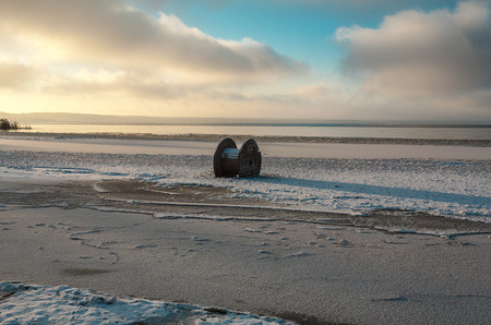 The empty coil from a high-voltage cable froze in ice near the coast.