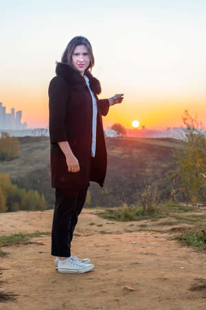 Young woman in a blue blouse and black coat at dawn, shows her finger in the sunの写真素材
