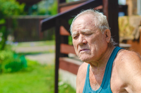 An elderly man in a green T-shirt sits at a country house with his eyes closedの写真素材
