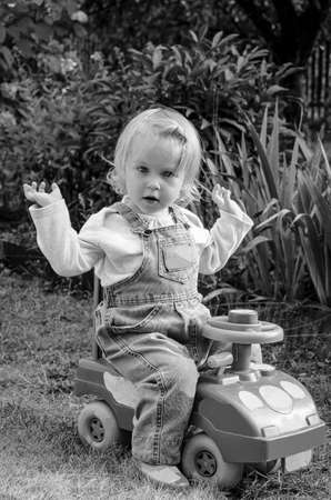 A little girl in denim overalls, driving dad's old car, in the summer garden.の写真素材