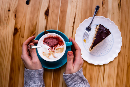 Female hands lie on the table and hold a cup of coffee on a wooden table, next to a piece of cakeの写真素材