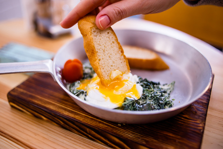 A woman dunks bread in scrambled eggs, breakfast consists of fried spinach, tomato, two slices of breadの写真素材