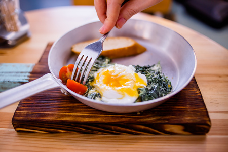 Hand fork pricks tomato, breakfast consists of fried spinach, scrambled eggs, two slices of breadの写真素材