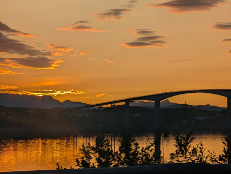 A captivating image of a bridge silhouetted against a breathtaking golden sunset in Norway. The sky...