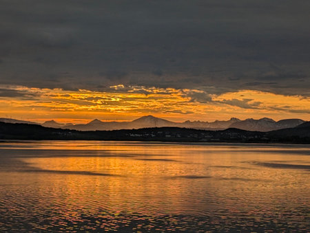 Vibrant orange sunset over calm fjord with golden water reflectionsの写真素材
