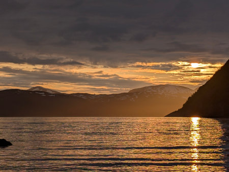 Golden sun setting behind snowy mountains with shimmering reflections on fjordの写真素材