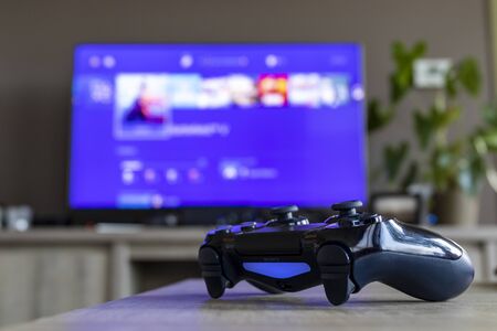 A portrait of a PS4 controller, which is turned on, in front of a television. The television is blurred out and displays the Playstation home screen.のeditorial素材