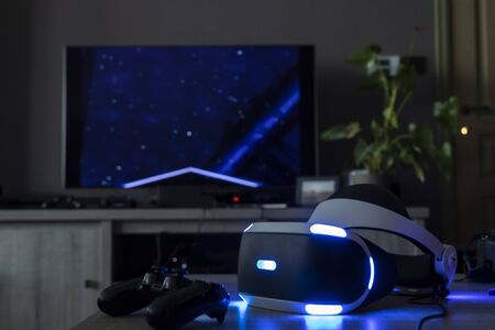 BRECHT,BELGIUM - 12 JUNE 2019: A Playstation VR headset lighting up the table next to a Playstation controller in a dark living room. Behind it is a television turned on with a game on it.のeditorial素材