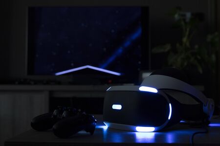 Brecht, Belgium - June 12 2019:  A playstation VR headset in a dark room showing its iconic lights in front of a television and a playstation 4 controller. Ready to play a game.のeditorial素材