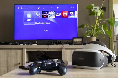 Brecht, Antwerp, Belgium - June 6 2019: A playstation 4 controller and some move controllers with a virtual reality headset on a table in front of a television showing the playstation home screen.のeditorial素材