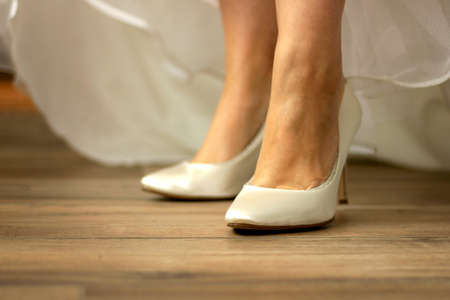 A portrait of the feet of a bride wearing white high heeled wedding shoes while pulling her dress up to show them. The woman is standing on a wooden floor with her high heels ready to get married.の写真素材