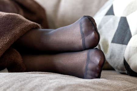 A close up portrait of the feet encased in pantyhose of a person lying in a couch under a brown blanket with a pillow in the back. The color of the nylon hosiery is black and the toes are reinforced.の写真素材