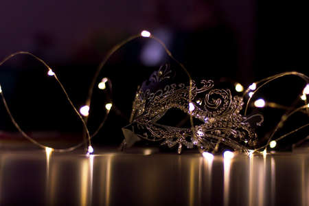 A scenic portrait of a mask lying on a table surrounded by fairy lights. The dark environment casts some mystery over the mask.の写真素材