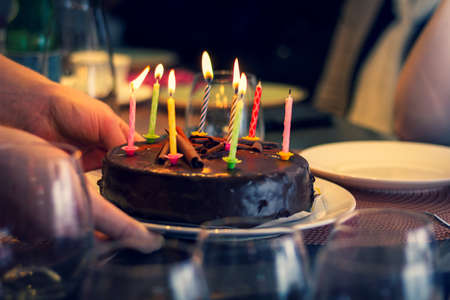 A portrait of a person putting a chocolate birthday cake with burning candles on it on a wooden festive table. Ready to be blown out and start eating and celebrating.の写真素材