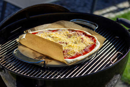 A portrait of a pizza margherita lying on a pizza stone and a piece of baking paper on a charcoal barbecue grill. The delicious pizza has cheese and tomato sauce on it.の写真素材