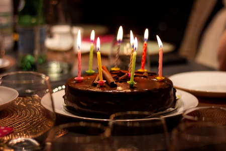 A portrait of a chocolate birthday cake with burning candles on it on a festive dining table. The lit candles all have different colors.の写真素材
