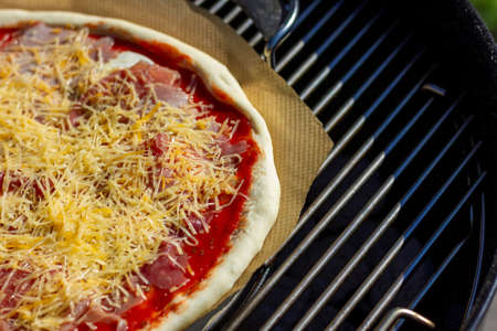 A closeup portrait of a pizza being baked on a piece of baking paper on a pizza stone on a barbecue grill. The pizza is topped with cheese, tomato sauce and other toppings.の写真素材