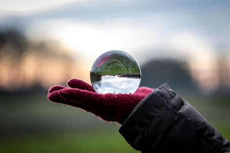 A scenic landscape image reflected inside of a lens ball resting on the red glove of a person. The landscape is a meadow during the start of sunset.の写真素材