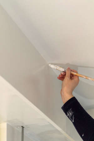 A portrait of a female hand and a dirty sleeve of a person holding a paint brush, painting in the corner made by the wall and the ceiling with white paint in a house renovation.の写真素材