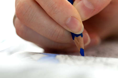 A close up portrait of a hand holding a blue colored pencil, whilst coloring in a book full of illustrations. A perfect way to relax from a hard day of work is with your hobby.の写真素材
