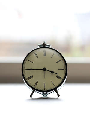 A portrait of a small old fashioned antique alarm clock with roman numbers on the dial on a very ligth background. The hands of the clock are almost pointing to a quarter to 4 o'clock.の写真素材