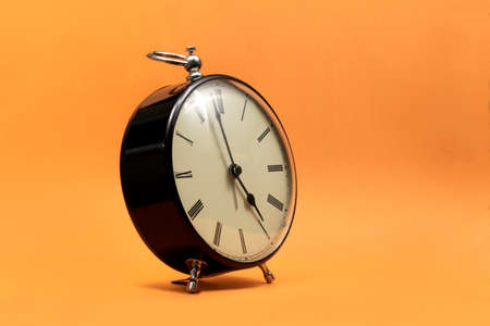 A portrait of a small old fashioned antique alarm clock with roman numbers on the dial on an orange background. The hands of the clock are almost pointing to 5 o'clock.の写真素材