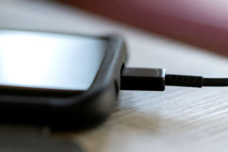 A close up portrait of a smartphone in a sturdy case charging on a wooden table with a USB cable in its port. The phone had to charge because its battery died.の写真素材