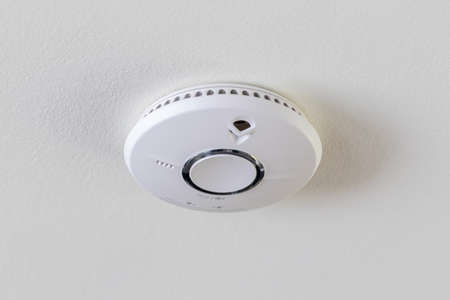 A close up portrait of a smoke detector hanging on a ceiling for safety. The device alerts people of a fire by making a sound. The fire alarm is round and hands on a white ceiling.の写真素材