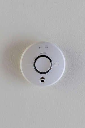 A close up portrait of a fire alarm sticking on a ceiling for safety. The device is round and hands on a white ceiling. The smoke detector alerts people of a fire by making a sound.の写真素材