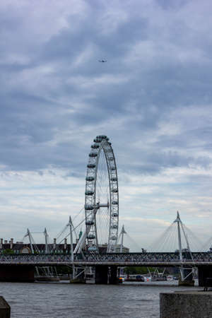 London / England - May 24 2019: A portrait of the London eye with a moody sky and an airplane above it and a bridge over the Thames.のeditorial素材