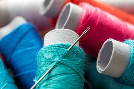 A portrait of a sewing needle stuck in a roll of thread. The spool of wire is lying on top of others rolls, all of them have a different color. The yarn or fiber is ready to be used in soem needleworkの写真素材