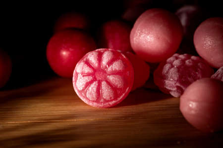 A dark portrait of some pieces of red and pink hard and round candy lying on a wooden table. The...