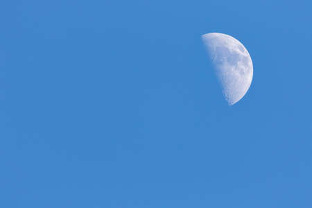 A portrait of a half moon during day time in a blue sky. The craters on the visible have of the satelite planet are well visible.の写真素材