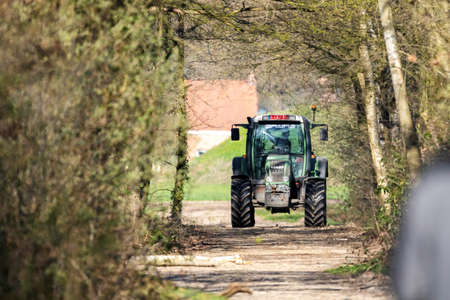 Brecht, Belgium - March 24 2019: A farmer driving a green Fendt tractor through a forest on a farm path. Both sides of the road have trees and the vehicle just passed the edge of the woods.のeditorial素材