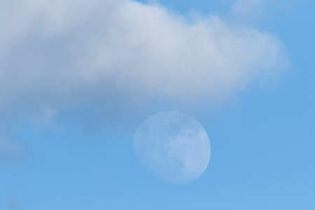 A portrait of a blue sky with a dark gray cloud in it and below the cloud the moon is faintly present.の写真素材