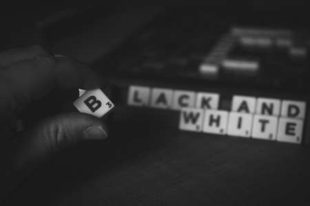 Brecht, Belgium - 28 July 2020: A hand of a person holding the letter b in between the fingers in front of a scrabble board with the words black and white on them in the blurry background.のeditorial素材
