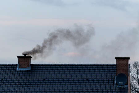 A roof with two chimneys on it. In one of the chimneys there is a pipe mounted on it were a lot of black polluting smoke is coming out or rising up from, connected to the fireplace of the house.の写真素材