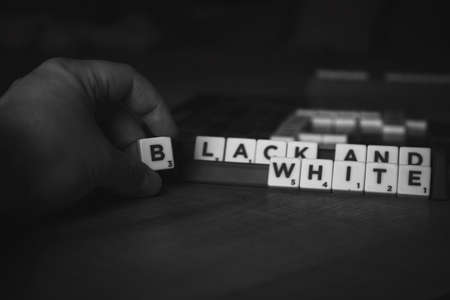 Brecht, Belgium - 28 July 2020: A black and white portrait of a hand of a person holding the letter b in between the fingers in front of a scrabble board with the words black and white on them.のeditorial素材