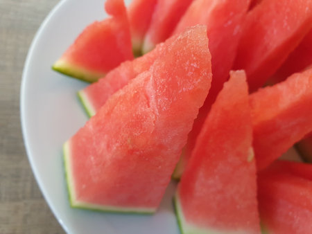 A close up portrait of red pointy slices of a cut watermelon on a white plate next to an uncut green, big and round one of the fruit. A great, healthy and refreshing snack on a hot summer day.の写真素材