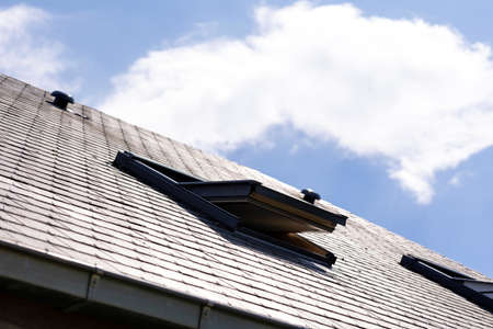 A portrait of an open skylight window for ventilation in a slate roof next to another one on a sunny day with a blue sky with white clouds. you can also see a ventilation vent on the rooftop.の写真素材