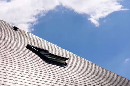 A portrait of an opened skylight window for ventilation in a slate roof on a sunny day with a blue sky with white clouds. you can also see a ventilation vent on the rooftop.の写真素材