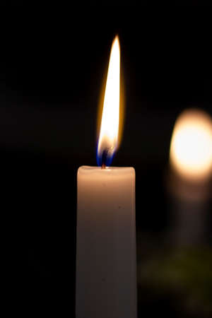 A close up shot of a lit thin white candle burning in the darkness, with a blurred one in the background. The flame is blue at the bottom and yellow at the top.の写真素材